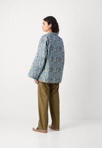 Giacca trapuntata blu con motivo floreale e maniche ampie, abbinata a pantaloni cargo verdi oliva e sneakers beige con dettagli neri.