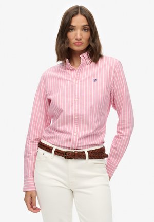 Camicia button-down a righe rosa e bianche con maniche lunghe, caratterizzata da un piccolo logo blu e indossata con una cintura intrecciata marrone sopra pantaloni bianchi.