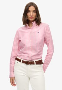 Donna che indossa una camicia a righe verticali rosa e bianche con bottoni e collo, pantaloni bianchi e una cintura intrecciata marrone, in piedi su sfondo bianco.
