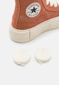 Zapatilla alta de gamuza naranja con suela de goma blanca y ojetes, que presenta un parche de estrella negra. Incluye dos cordones blancos.