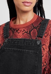 Svarta denimoveraller med justerbara remmar. Kombinerat med en långärmad skjorta i röd ormskinnsmönster. Fokusera på textur och färgkontrast.