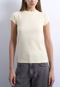 Γυναίκα που φοράει κοντομάνικη εφαρμοστή μπλούζα ανοιχτού κίτρινου χρώματος και σκούρο γκρι τζιν παντελόνι, μπροστά από απλό φωτεινό τοίχο.