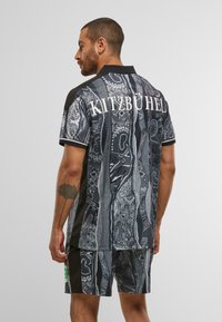 Mann mit kurzen Haaren und Tattoo, der schwarze und graue gemusterte Shorts sowie ein passendes Shirt mit dem Aufdruck "KITZBÜHEL" auf dem Rücken trägt.