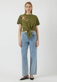 T-shirt verde oliva con ricamo floreale, annodato in vita. Abbinato a jeans in denim azzurro chiaro a gamba larga e ballerine nere a punta.