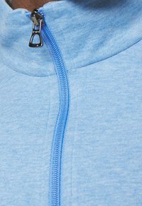 Ljusblå fleecejacka med dragkedja och hög krage, med en kontrasterande blå dragkedja och texturerat, mjukt tyg. Inga synliga mönster eller logotyper.