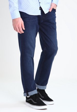 Homme portant un jean bleu foncé retroussé, des baskets noires avec des semelles blanches et une chemise bleu clair à petits motifs en points.