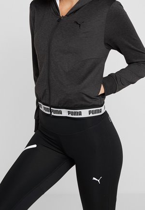 Kobieta ubrana w czarną bluzę z kapturem Puma zapinaną na suwak oraz czarne legginsy Puma, z rękami w kieszeniach, na jednolitym szarym tle.
