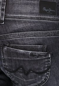 Jeans de mezclilla gris oscuro con costuras prominentes, un bolsillo trasero con un diseño curvado y una etiqueta de cuero negra con "Pepe Jeans London".