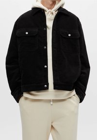 Giubbotto in corduroy nero con colletto, due tasche sul petto e bottoni a pressione argentati. Indossato sopra un hoodie crema e pantaloni chiari.