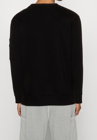 Felpa nera con maniche lunghe e scollatura rotonda. Presenta una tasca a toppa sulla manica sinistra. Tessuto morbido e testurizzato.