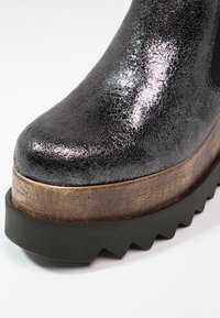 Mustan kiiltävä halkeillut nahkainen platform-kengät, joissa on puinen välikerros ja paksu musta sahalaitainen kumipohja.