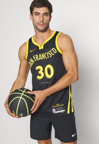 Camiseta de baloncesto negra con acentos amarillos, que presenta "SAN FRANCISCO" y el número "30". El modelo sostiene un balón de baloncesto negro y verde, vistiendo pantalones cortos negros.
