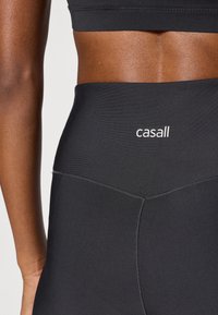 Leggings neri a vita alta con tessuto liscio ed elastico. Presenta un logo "casall" bianco sulla fascia in vita e dettagli di cuciture sottili.