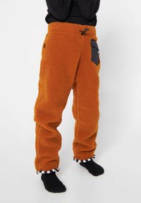 Orange fleecebukser med sorte nylonlommer og snøre i taljen; har stribet stof ved anklerne. Blød tekstur, afslappet pasform.