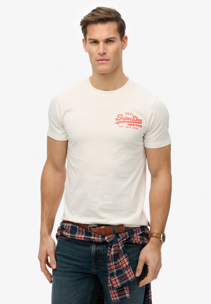 Camiseta blanca de algodón con el logo rojo de "SuperDry". Combinada con jeans de mezclilla oscura y una camisa de cuadros atada a la cintura.