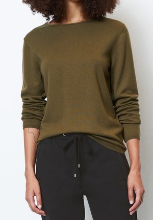 Personne portant un pull vert olive à manches longues et un pantalon noir à cordon, avec un tatouage visible sur l'avant-bras gauche.