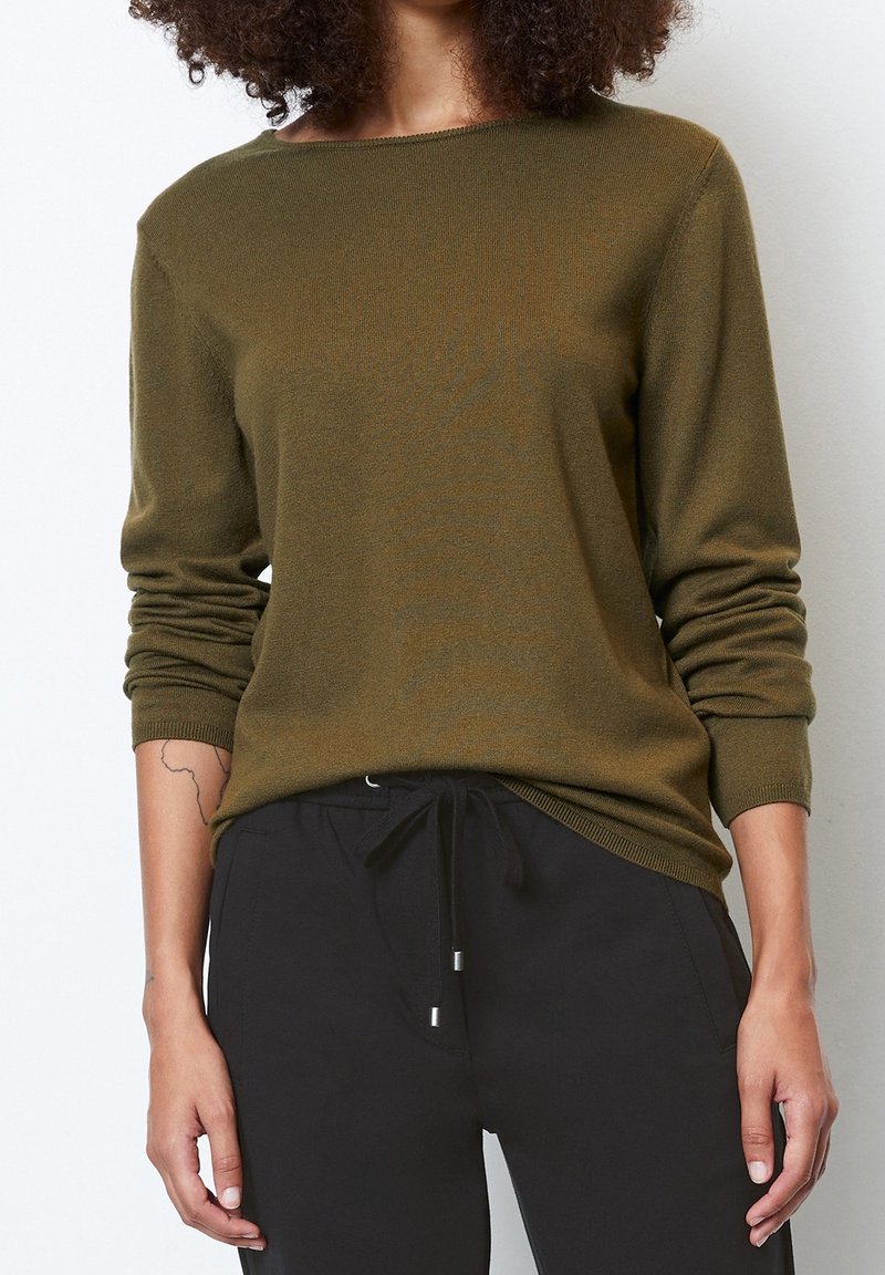 Personne portant un pull vert olive à manches longues et un pantalon noir à cordon, avec un tatouage visible sur l'avant-bras gauche.