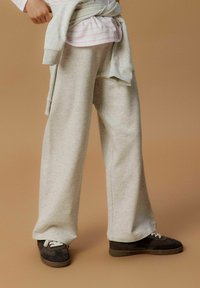 Pantalons de survêtement larges gris clair avec une texture douce. Associés à des baskets sombres avec des lacets blancs. L'arrière-plan est beige neutre.