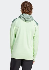 Sudadera de manga larga verde claro con tejido texturizado, capucha de malla y detalles en verde más oscuro en los hombros. Corte relajado y dobladillo ligeramente curvado.
