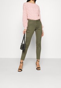 Pull en tricot rose avec un motif texturé, associé à un pantalon cargo vert olive avec des poches latérales. Sac à main noir et talons à brides.