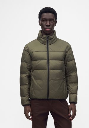 Mann trägt eine olivgrüne Steppjacke mit hohem Kragen und steht vor einem schlichten weißen Hintergrund.