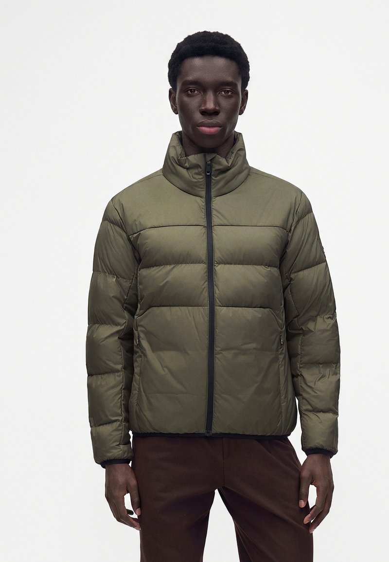 Homme portant une doudoune vert olive à col montant, debout devant un fond blanc uni.