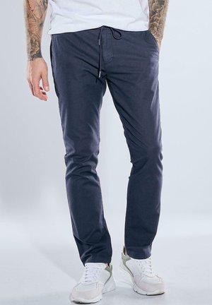 Mann trägt dunkelblaue Jogginghose mit Kordel und weiße Sneaker, steht mit einer Hand in der Tasche vor einfarbigem Hintergrund.