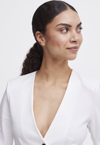 Hvid beklædning med dyb V-hals og glat tekstur samt lange ærmer, der har en enkelt sort knap i taljen. Hudtonen er neutral.