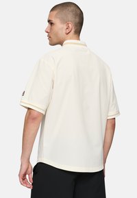 Polo a maniche corte in bianco sporco, realizzato in tessuto texturizzato con dettagli a righe a contrasto nel collo e nei polsini. Indossato, mostra la vista posteriore.