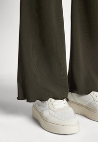 Kaki ribbede vide bukser med bølget kant, kombinert med hvite joggesko med tekstur og perforerte detaljer.