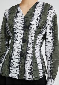 Blouse verte et blanche à motifs avec un décolleté en V, trois boutons et un léger froncé à la taille, ornée de larges rayures noires semblables à l'écorce d'arbre.