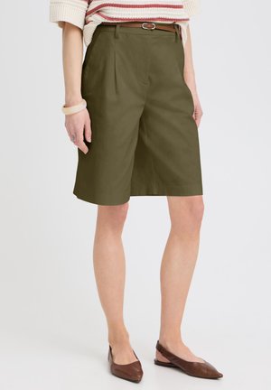 Naine, kes kannab oliivrohelisi põlveni ulatuvaid lühikesi pükse, kitsast pruuni vööd, triibulist kampsunit, beež käevõru ja pruune teravnurkseid jalanõusid.