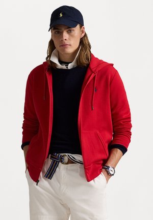 Rote Zip-Hoodie mit Kapuze und Fronttaschen, über einem dunklen Pullover und einem Hemd mit Kragen getragen. Kombiniert mit weißen Hosen und einem gestreiften Gürtel.