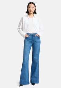Weißes Button-Down-Hemd über einem eng geschnittenen weißen Oberteil, kombiniert mit hochgeschnittenen, ausgestellt geschnittenen blauen Jeans und schwarzen spitzen Pumps.