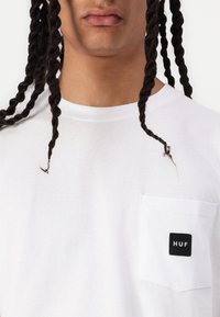 Tricou alb cu un buzunar pe pieptul stâng, având un petic pătrat negru cu "HUF" scris în text alb. Textură netedă, guler de tip crew.