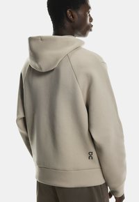 Sweatshirt beige à capuche en tissu texturé, présentant une coupe décontractée, des poignets côtelés et un accent logo dans le bas du dos.