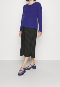 Maglione viola a V, gonna midi nera plissettata e sandali blu con piattaforma e multiple cinghie. L'outfit combina elementi casual e formali.