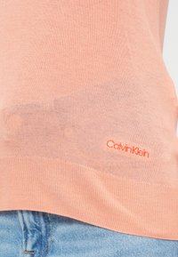 Breskvaste barve pulover Calvin Klein z gladko teksturo, rebrastim robom in izvezenim logotipom v kontrastni oranžni barvi na dnu.