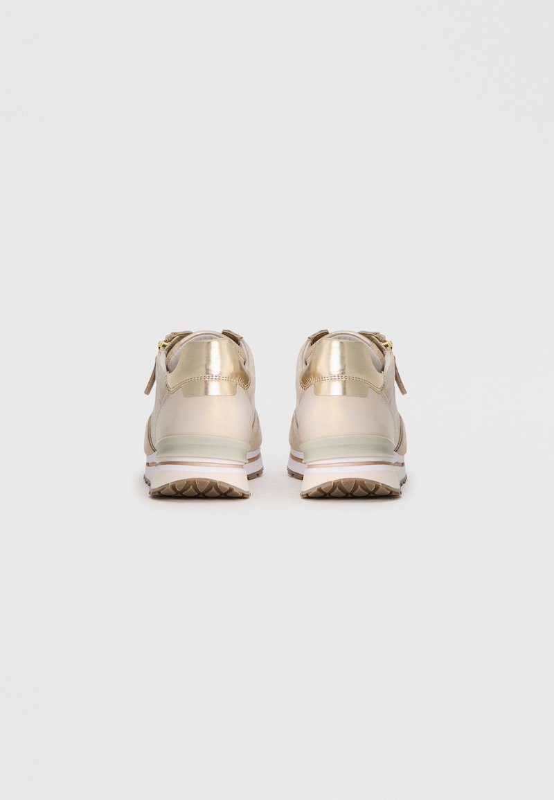 Helle beige Sneakers mit metallisch goldenen Akzenten, die eine glatte Oberfläche, eine dicke weiße Sohle und eine strukturierte braune Außensohle aufweisen. Reißverschlussdetail auf der Rückseite.