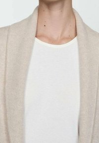 Beige neuletakki, joka on valmistettu pehmeästä kankaasta. Siinä on avoin etuosa ja huivikaulus, joka on kerroksittain yllään yksivärisen valkoisen, ribbineuloksen tekstuurilla varustetun t-paidan päällä.