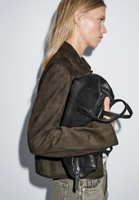 Black leather bag with soft texture, carried in a hand. The person wears a brown suede jacket with a cropped design and light-colored jeans.