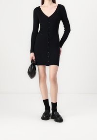 Robe noire côtelée à manches longues avec un décolleté en V et une fermeture à boutons à l'avant. Associée à des mocassins noirs et un petit sac à main texturé.