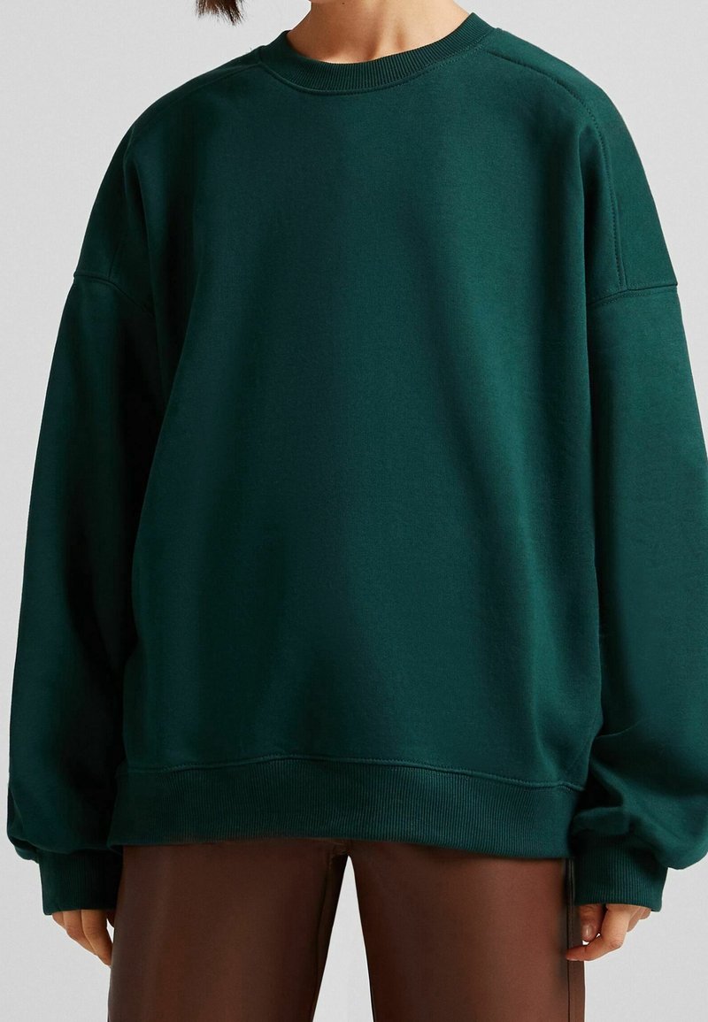 Personne portant un sweat-shirt oversize vert foncé à col rond et un pantalon marron, vue du cou jusqu'à mi-cuisse sur un fond uni.