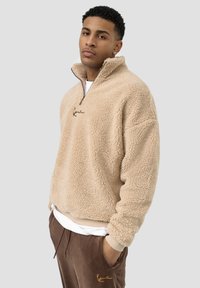 Beige fleece quarter-zip genser, med en myk, teksturert overflate, med brodert logo og kontrasterende krage. Kombinert med brune joggebukser.