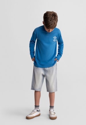 Chłopiec w niebieskiej koszulce z długim rękawem, jasnoszarych spodenkach do kolan, szarych skarpetkach i białych tenisówkach, patrzący w dół z rękami w kieszeniach.