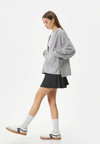 Grå hættetrøje med oversize ærmer og forlommer, båret over en sort plisséet nederdel og parret med hvide sneakers og knælange sokker.