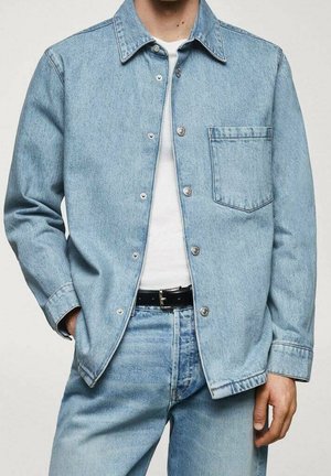 Homme portant une veste en jean bleu clair sur une chemise blanche, associée à un jean en jean bleu clair assorti et une ceinture noire.