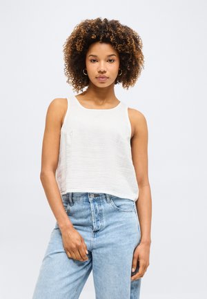 Γυναίκα με σγουρά μαλλιά που φοράει κρίκους στα αυτιά, αμάνικη λευκή μπλούζα και ανοιχτό μπλε τζιν με ψηλή μέση, στέκεται μπροστά σε απλό φόντο.