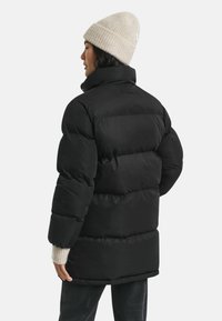 Schwarze Daunenjacke mit horizontalen Steppnähten, hohem Kragen und geripptem beigefarbenem Beanie; warmes, strukturiertes Material und lässige Passform.