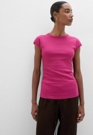 Vrouw draagt een aansluitend roze shirt met korte mouwen en een donkere broek, staat tegen een witte muur en kijkt naar de zijkant.